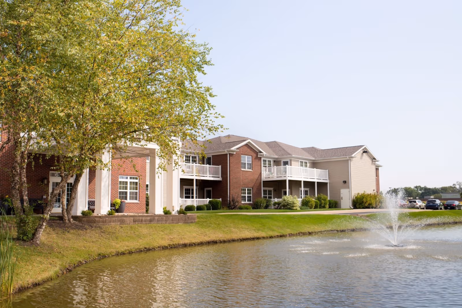 Exterior view of a senior living facility with a pond and a water fountain in the foreground, a tree on the left, and a two-story building with balconies and brick and siding facade in the background under a clear sky.