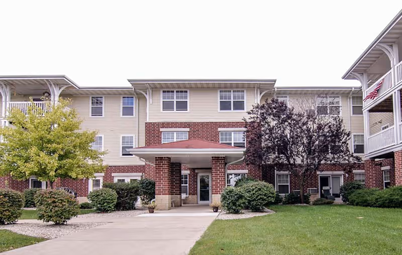 Front exterior view of Bell Tower Place Senior Apartments, a three-story building with a covered entrance supported by brick columns. The building features a combination of red brick and beige siding, surrounded by green grass, bushes, and trees.