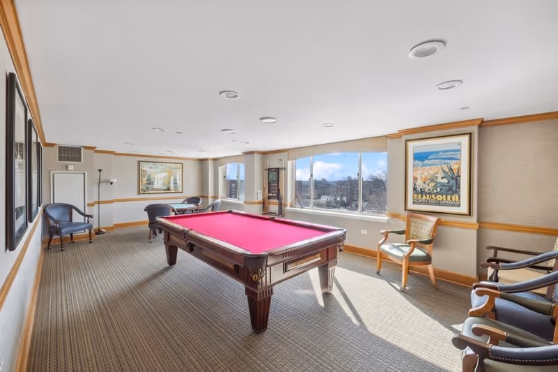 A bright recreational room with a red felt pool table in the center, surrounded by several chairs. Large windows on one side let in natural light and offer a view of trees and buildings outside. The walls are decorated with framed artwork, and the room has beige carpeting and light-colored walls with wooden trim.