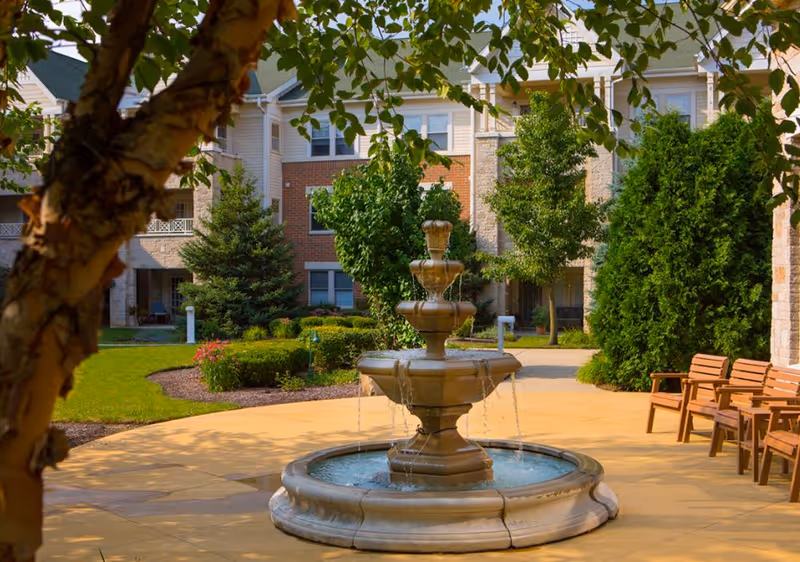 Tiered stone fountain in a landscaped courtyard with benches and a multi-story residential building behind.