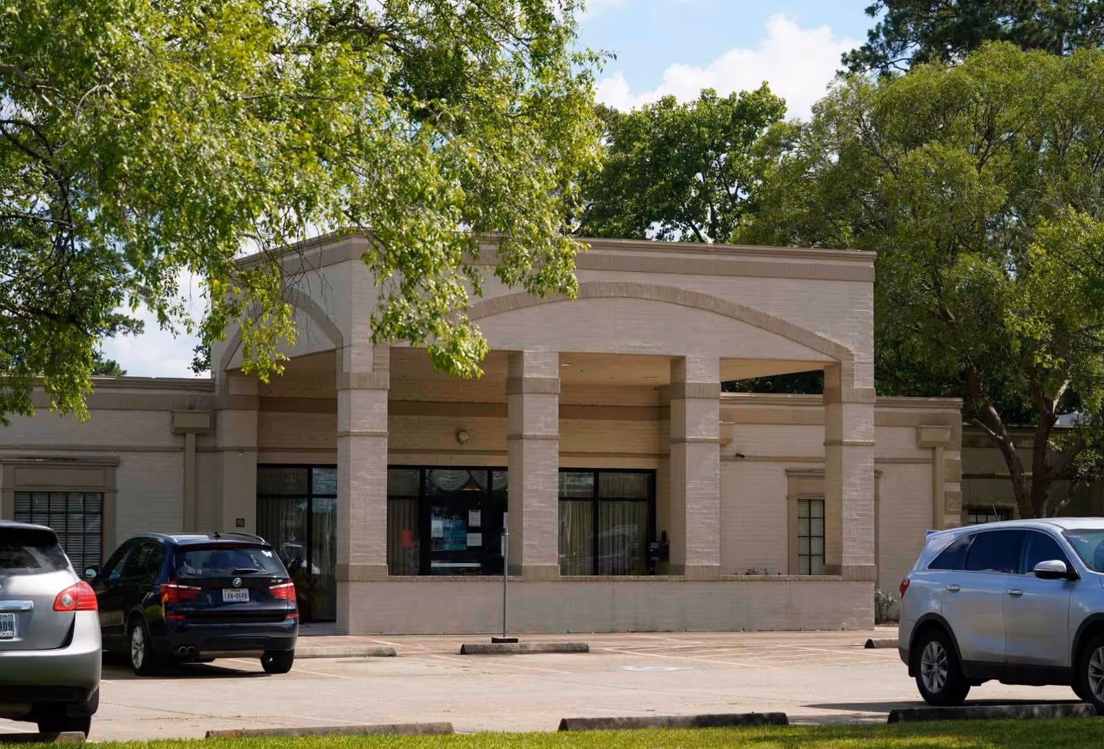 Exterior view of Oakmont Healthcare & Rehabilitation Center of Humble showing the front entrance with large pillars and a covered entryway. Several cars are parked in front, and trees with green leaves surround the building.