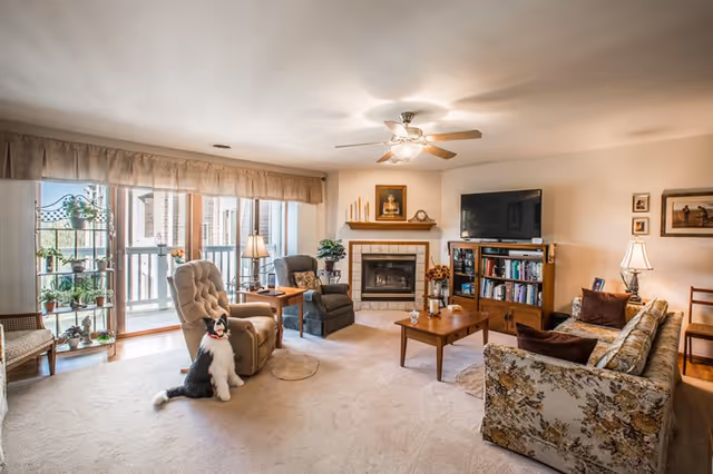 A cozy living room with a beige carpet, floral patterned sofa, two recliner chairs, a wooden coffee table, and a fireplace with a portrait above it. There is a flat-screen TV on a wooden stand with bookshelves, lamps on side tables, and a sliding glass door leading to a balcony with plants on a metal shelf. A black and white dog is sitting on the carpet near one of the recliners.
