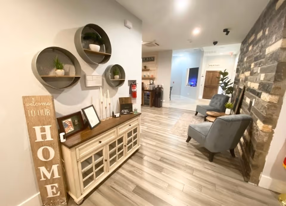 Interior view of a cozy common area with light wood flooring, a stone accent wall, two gray upholstered chairs, a wooden side table with a plant, and a cream-colored cabinet with glass doors. Above the cabinet are three round wall shelves with small plants and decor. A wooden sign leaning against the wall reads 'welcome TO OUR HOME'. In the background, there is a pantry door and a small bar area with a coffee machine and wine cooler.