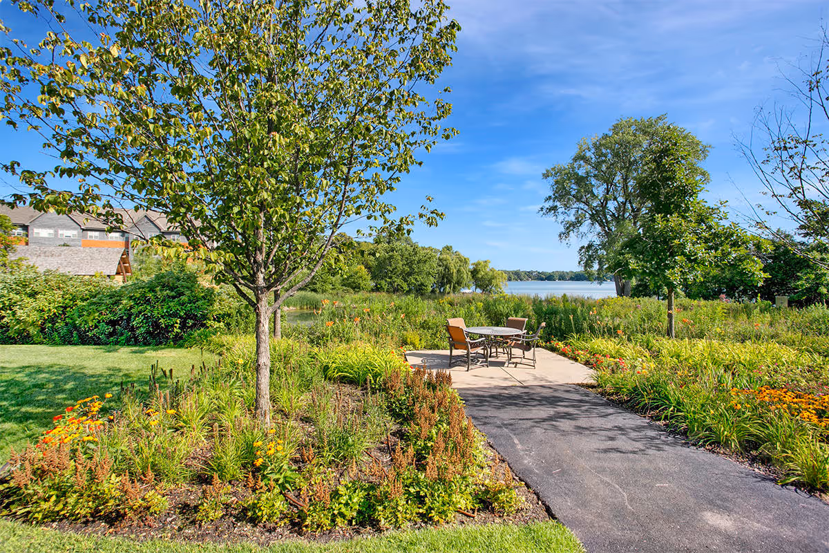 A scenic outdoor garden area with a paved pathway leading to a small patio with a table and four chairs. The garden is lush with green plants, colorful flowers, and several trees. In the background, there is a view of a lake under a clear blue sky.