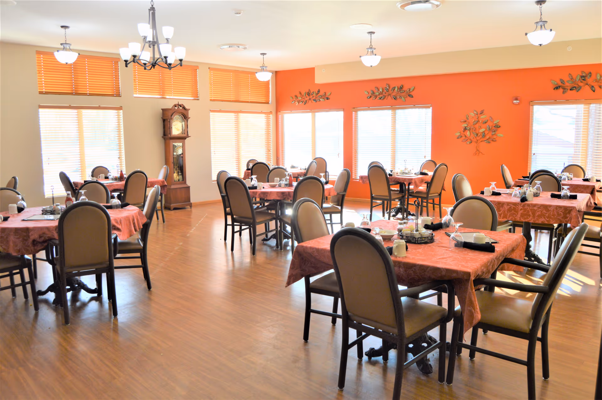 A bright dining room with multiple tables covered in red tablecloths, each set with chairs, glassware, and condiments. The room has large windows with blinds, an orange accent wall decorated with metal tree art, wooden flooring, and ceiling light fixtures including chandeliers.