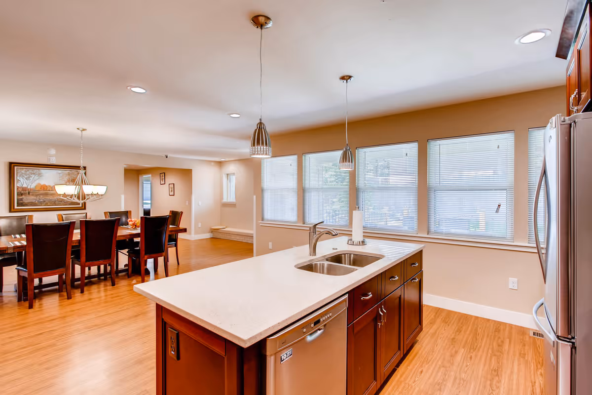 Bright and spacious kitchen area with a large island featuring a double sink and dishwasher. The kitchen has wooden cabinets and stainless steel refrigerator. In the background, there is a dining area with a wooden dining table and chairs, a chandelier, and a framed painting on the wall. Multiple windows with blinds allow natural light to fill the space.