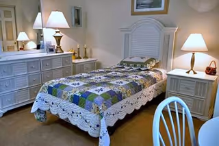 A small bedroom with a single bed covered by a patchwork quilt, white nightstands and dresser with lamps and a mirror.