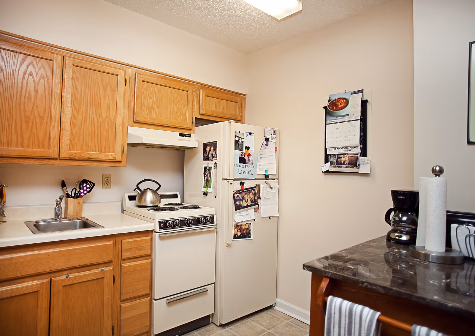 Compact kitchen with oak cabinets, a white stove and refrigerator covered in photos and magnets, a sink, and a countertop holding a coffee maker and paper towels.