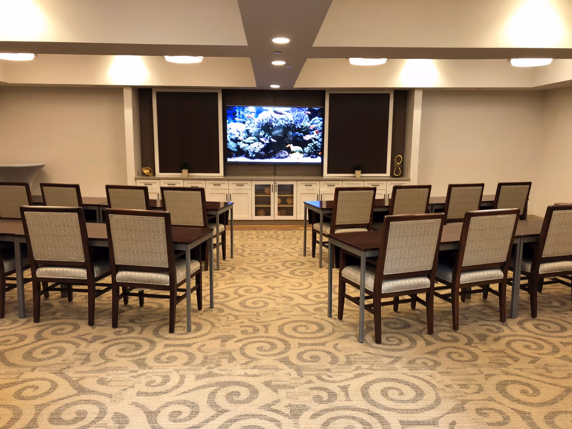 Meeting room with rows of chairs around tables facing a wall-mounted TV.