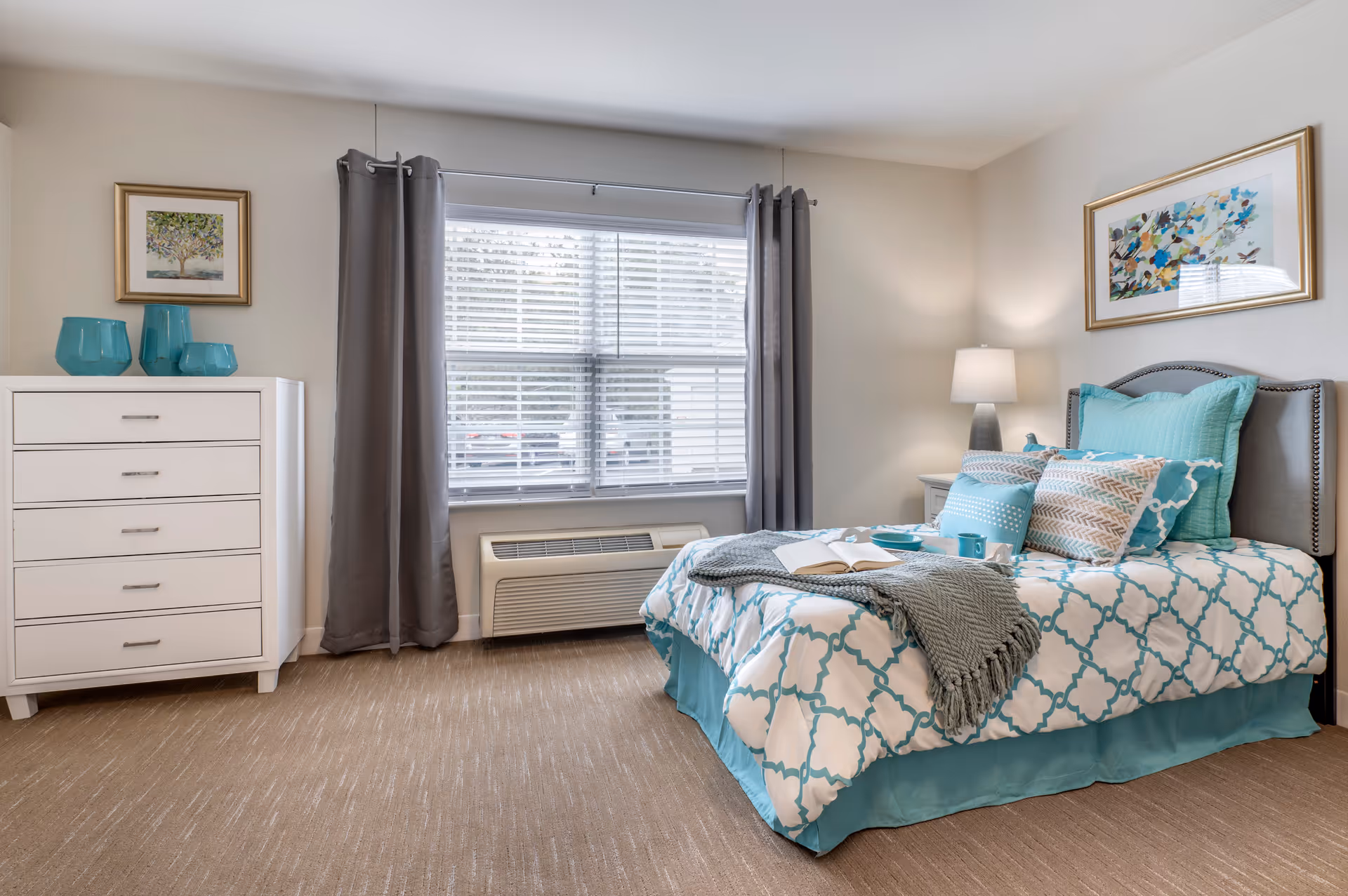 A neatly arranged bedroom with a teal-and-white patterned bed, nightstand with lamp, white dresser, and a window with gray curtains.
