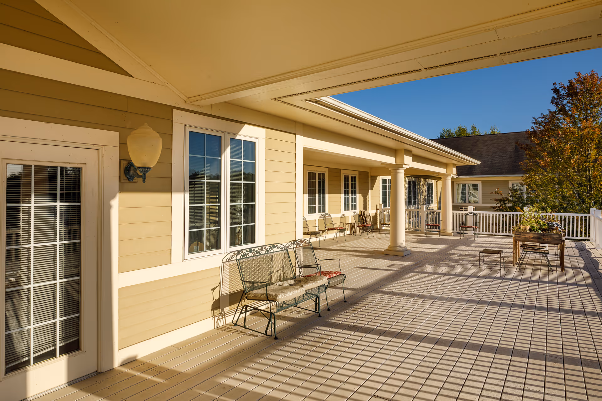A spacious outdoor patio area at Homestead Assisted Living & Memory Care of Centerville featuring a beige building exterior with white trim, several windows, a door with glass panes, metal benches with cushions, a table with chairs, and a planter box. The patio is covered partially by a roof supported by columns and is bathed in sunlight with clear blue skies and some trees in the background.