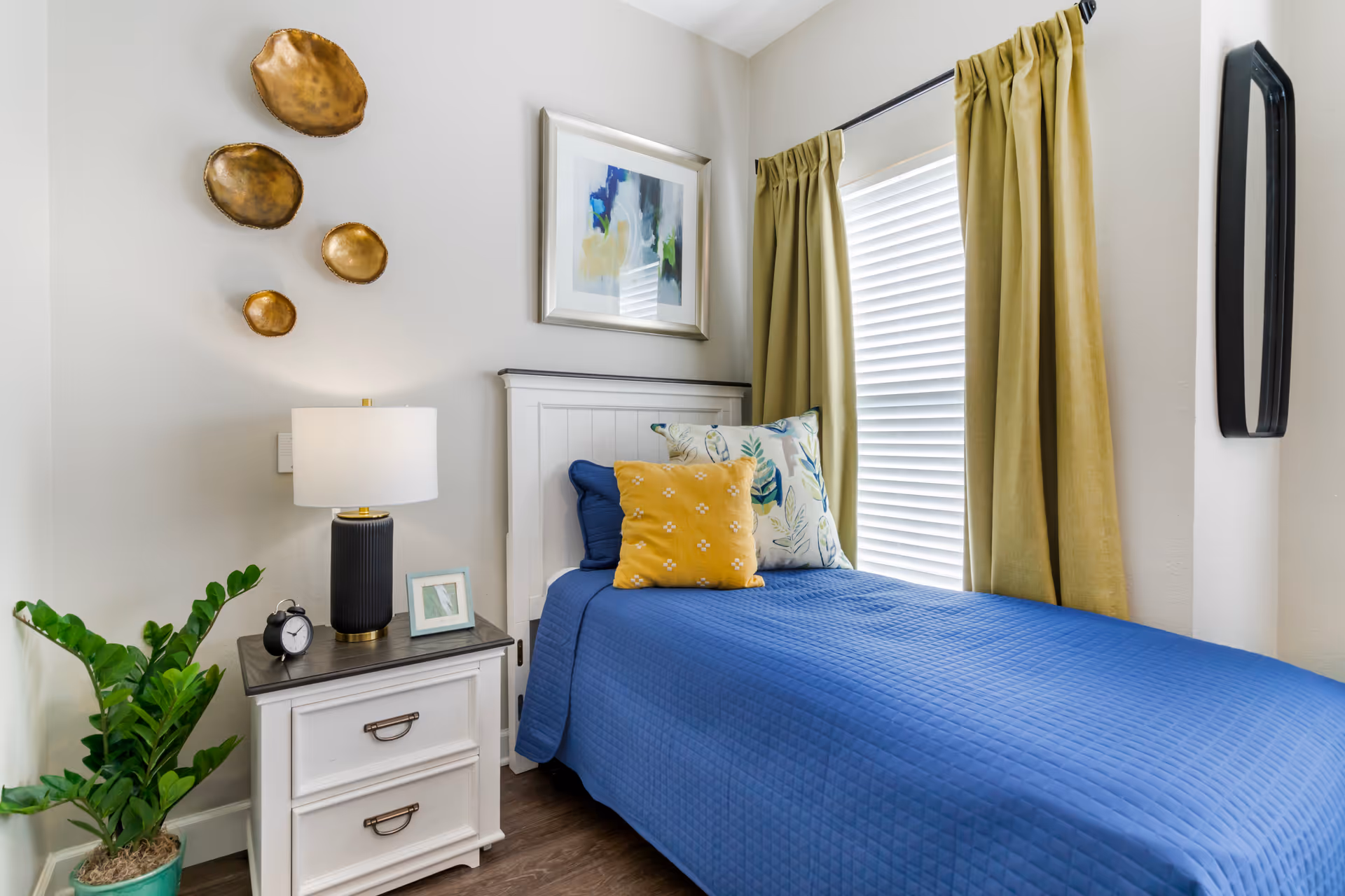 A cozy bedroom with a single bed covered in a blue quilt and decorated with three pillows in yellow and floral patterns. Next to the bed is a white nightstand with two drawers, holding a black table lamp, a small black alarm clock, and a framed photo. A green potted plant is placed on the floor beside the nightstand. The walls are light-colored and decorated with four gold decorative plates and a framed abstract painting. A window with closed blinds and mustard yellow curtains is beside the bed.