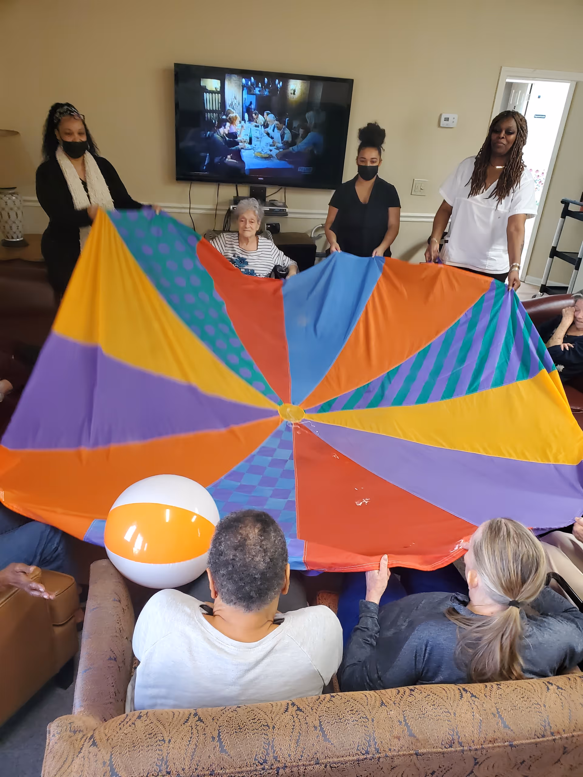 Several residents and staff in a common lounge hold a large colorful parachute with a beach ball.