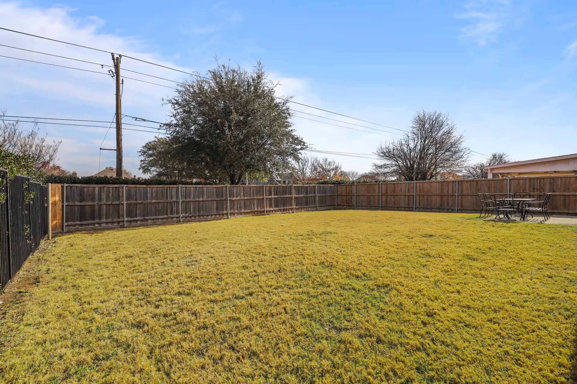 A fenced outdoor grassy yard with a few trees and a patio area with a table and chairs under a clear blue sky.