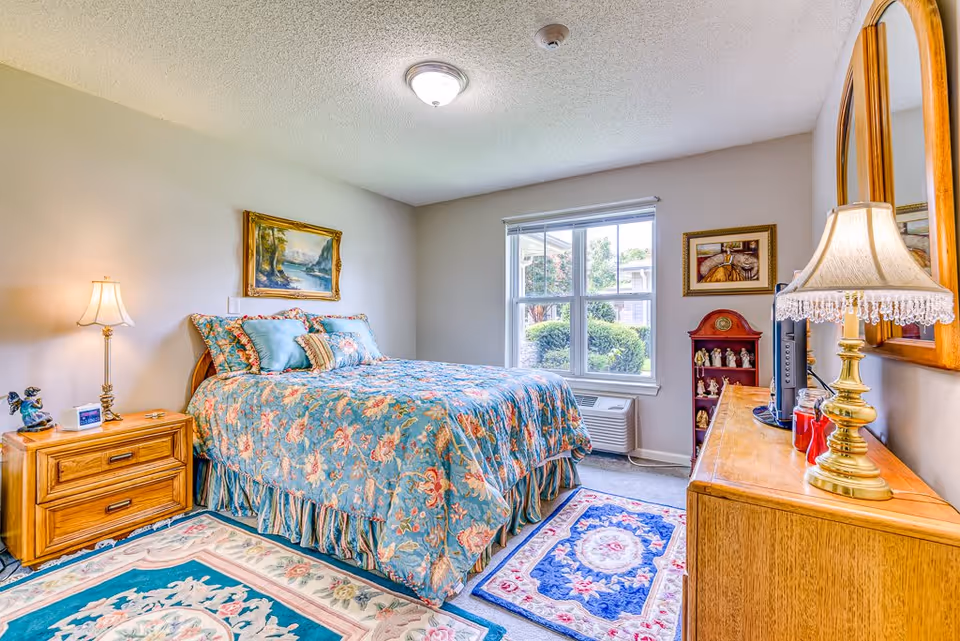 A cozy bedroom in Chesapeake Place Senior Living featuring a bed with floral bedding and multiple pillows, a wooden nightstand with a lamp and clock, a dresser with a decorative lamp and mirror, a window with a view of greenery outside, and framed artwork on the walls.