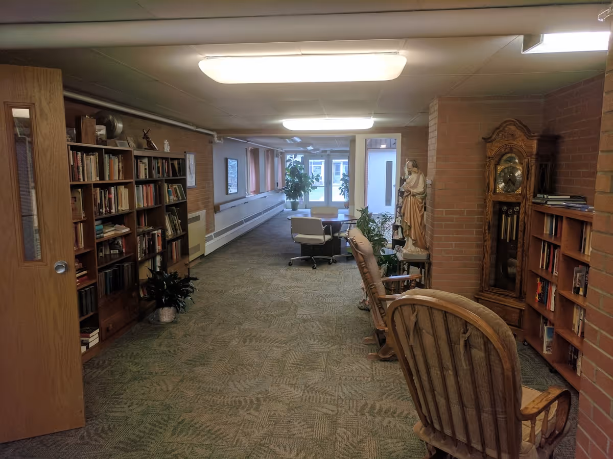 Interior hallway of St. Ann's Home featuring bookshelves filled with books on both sides, a wooden rocking chair, a grandfather clock, a statue, several potted plants, and a table with chairs near the far end by windows and a door.