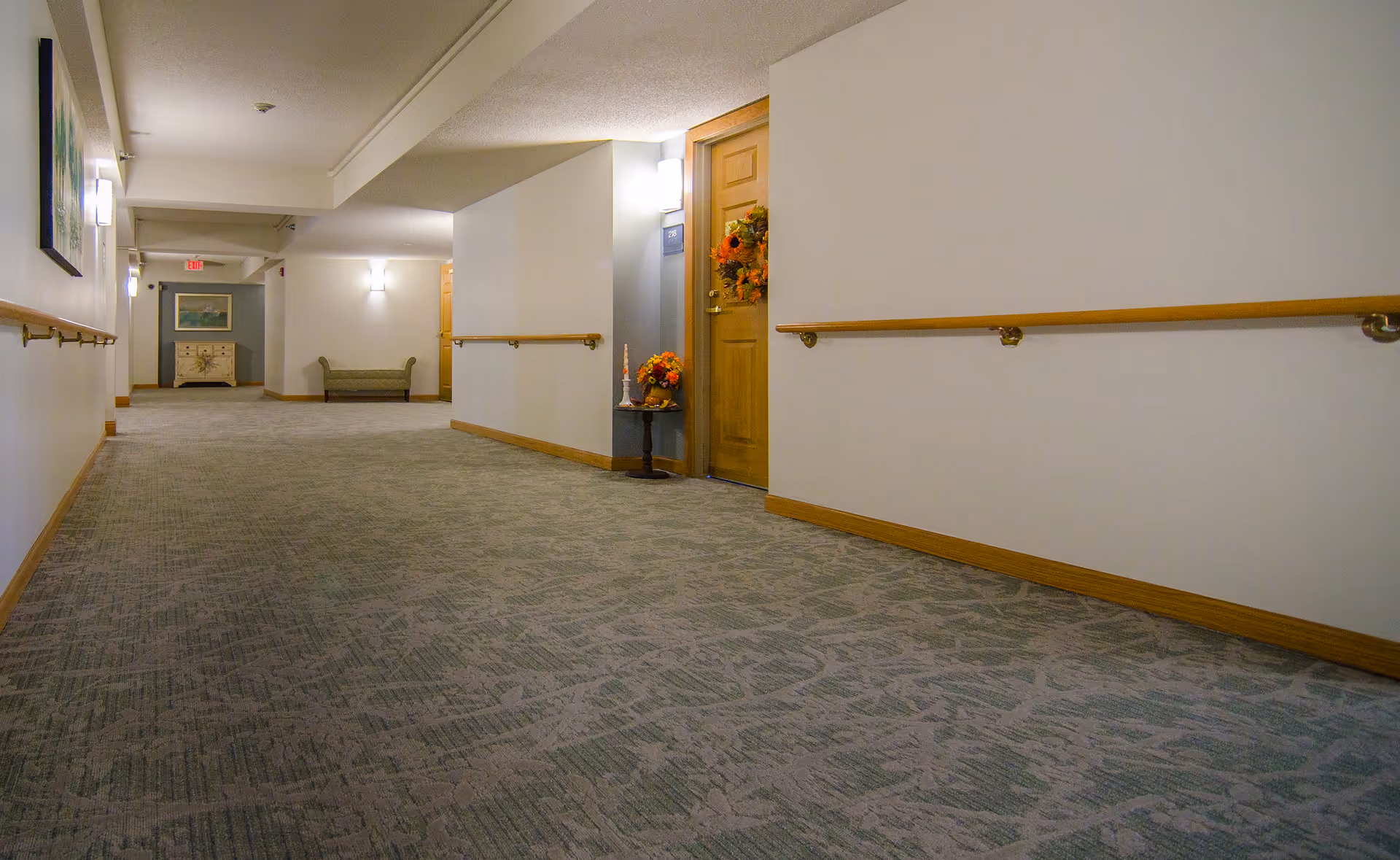 Carpeted interior hallway with wooden handrails, a decorated apartment door, and seating at the far end.