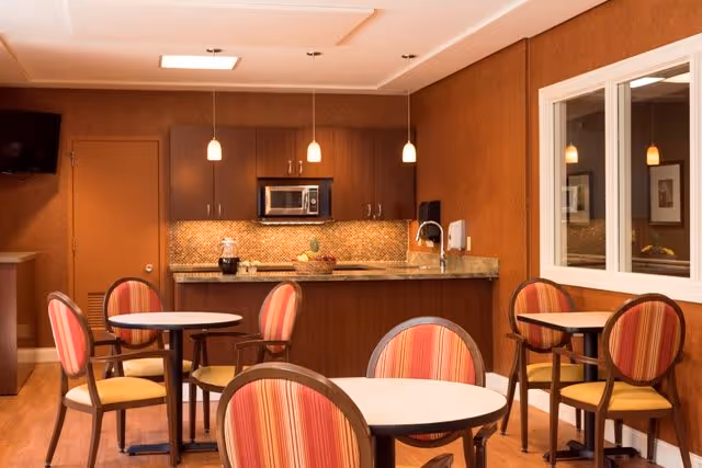 Dining area with round tables and striped chairs in front of a kitchenette with pendant lights and a microwave.