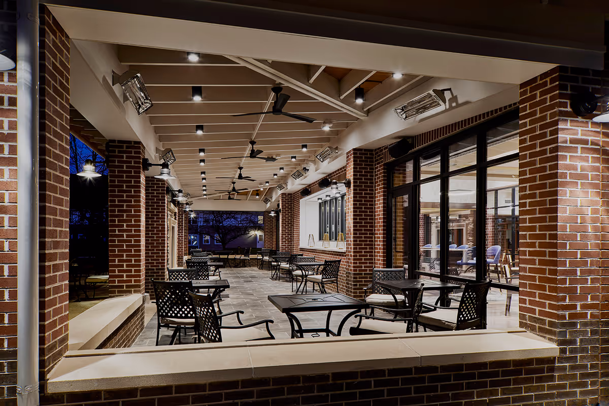 Covered outdoor patio area at night with brick pillars, ceiling fans, and multiple tables and chairs arranged for seating. Large windows on one side show an indoor seating area with armchairs.