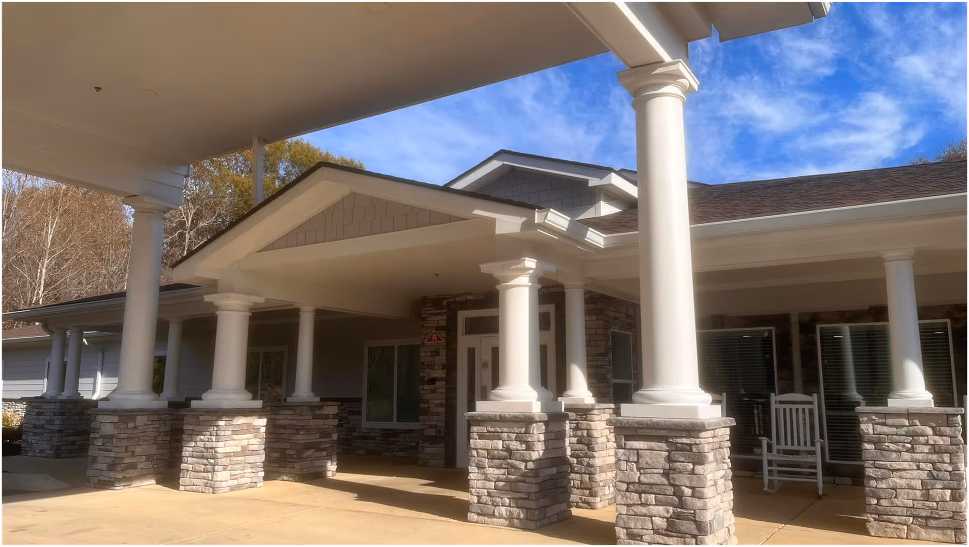 Front exterior view of a building with white columns on stone bases supporting a covered entrance. There are rocking chairs on the porch and trees in the background under a blue sky with some clouds.