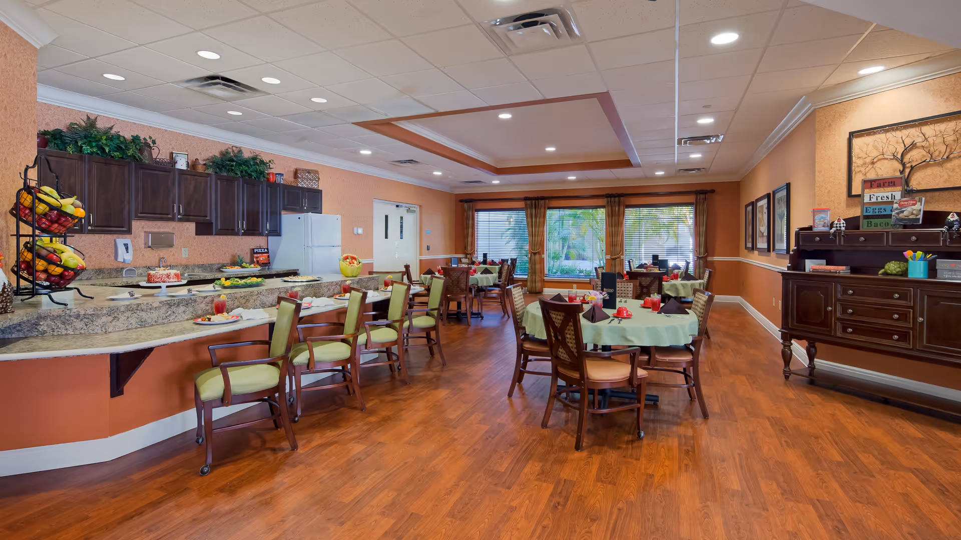 A spacious dining room with wooden floors and orange walls. Several round tables are set with green tablecloths, red cups, and napkins. There are wooden chairs around the tables and a counter with green cushioned chairs. The counter has fruit baskets and plates with food. Large windows with brown curtains let in natural light. A dark wooden sideboard with decorative items and framed pictures on the wall is visible on the right side.