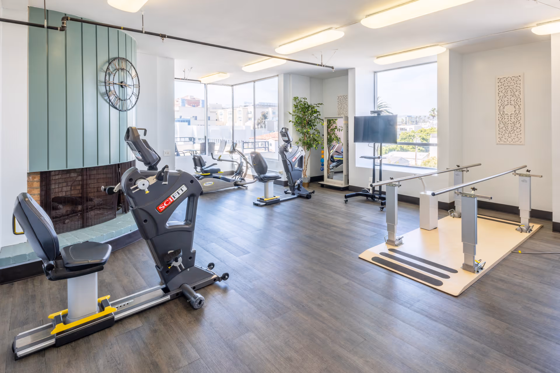 A bright and spacious rehabilitation exercise room with large windows letting in natural light. The room features several pieces of exercise equipment including a recumbent bike, an elliptical machine, and parallel bars for walking practice. There is a large wall clock above a fireplace with a green tiled surround, a full-length mirror, a TV on a stand, and a potted plant near the windows.
