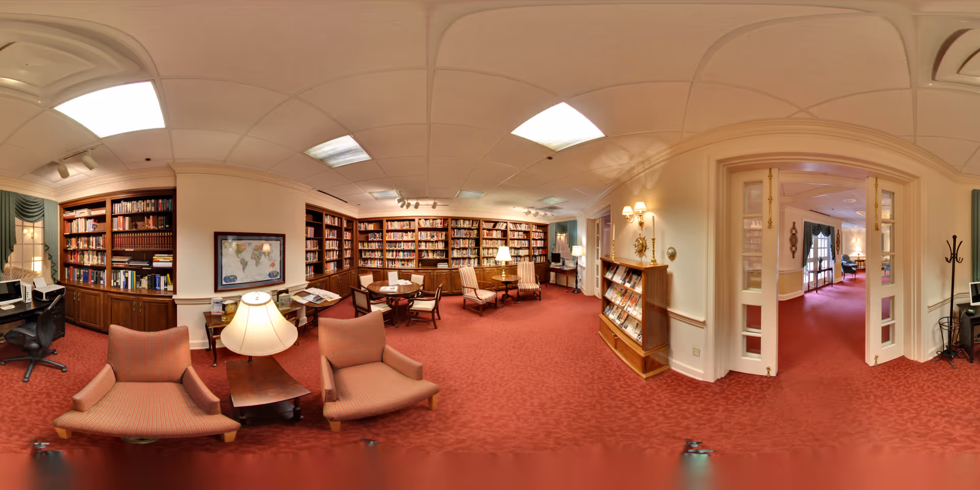 A spacious library room with red carpet flooring and cream-colored walls. The room features multiple wooden bookshelves filled with books, several comfortable upholstered chairs, a round table with chairs, and a lamp on a side table. There is a framed world map on one wall and a magazine rack near a doorway leading to another room with similar decor. The ceiling has recessed lighting and fluorescent panels.