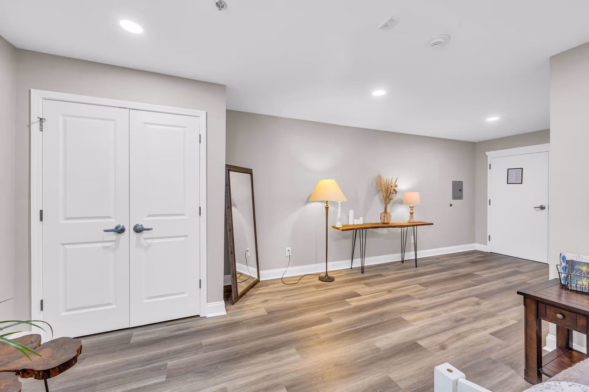 A spacious interior room with light gray walls and wood flooring. There is a double white door on the left, a full-length mirror leaning against the wall, and a wooden console table with hairpin legs holding two lamps and decorative items. A white door with a paper notice is visible on the right side of the room.