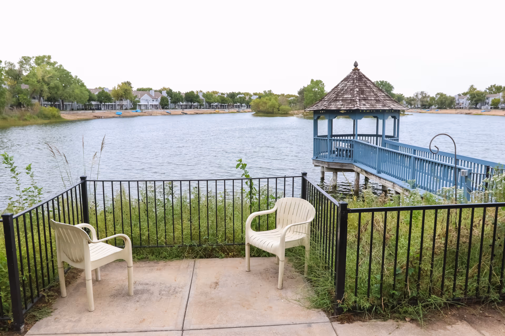 Two white plastic chairs on a concrete patio facing a lake with a blue wooden gazebo on stilts connected by a walkway. Trees and houses are visible across the lake.