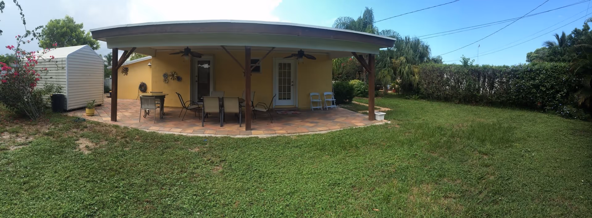 A backyard patio area with a covered seating space featuring a table and several chairs. The patio has a tiled floor and ceiling fans. There is a yellow building wall with two doors leading inside. The yard has green grass, some bushes, palm trees, and a white shed on the left side.