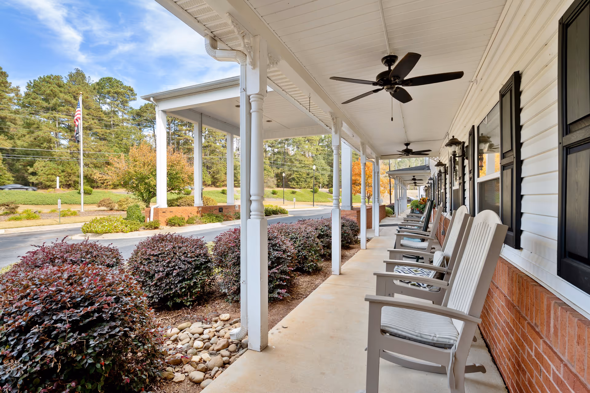 A covered outdoor porch area at Highlands Senior Living Jefferson with white rocking chairs lined up along the wall, ceiling fans, and decorative bushes along the walkway. Trees and an American flag are visible in the background.