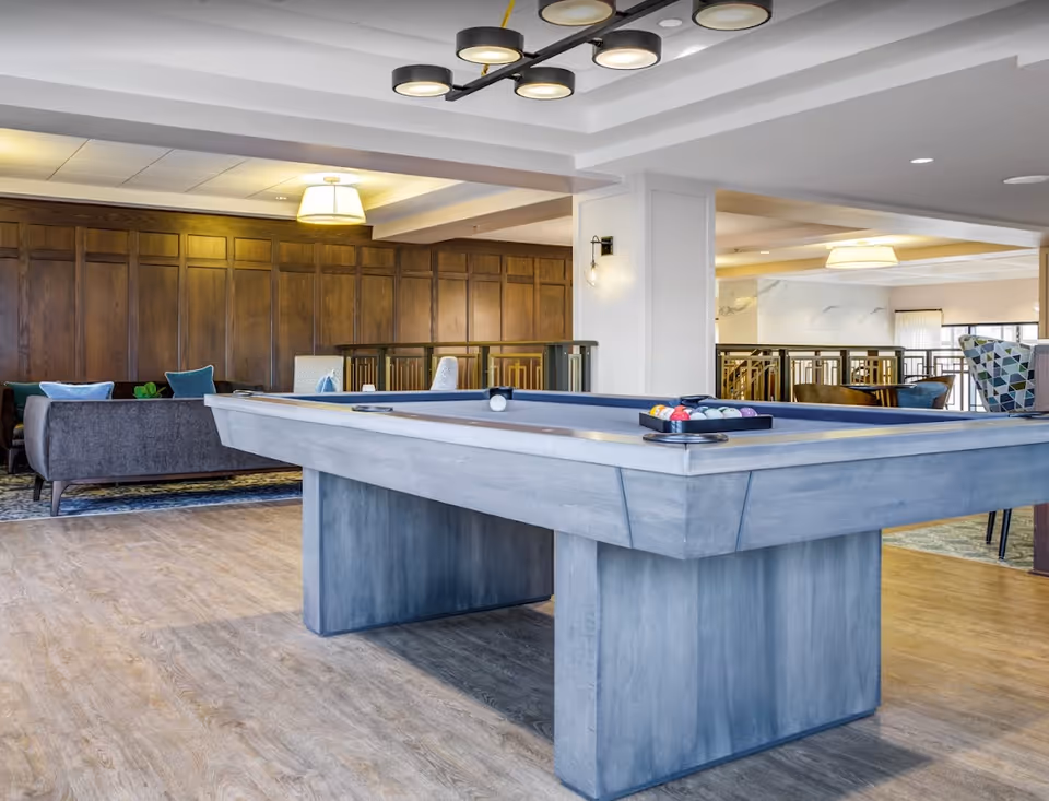 Modern communal game room with a gray pool table in the foreground and seating area with sofas and wood-paneled wall in the background.