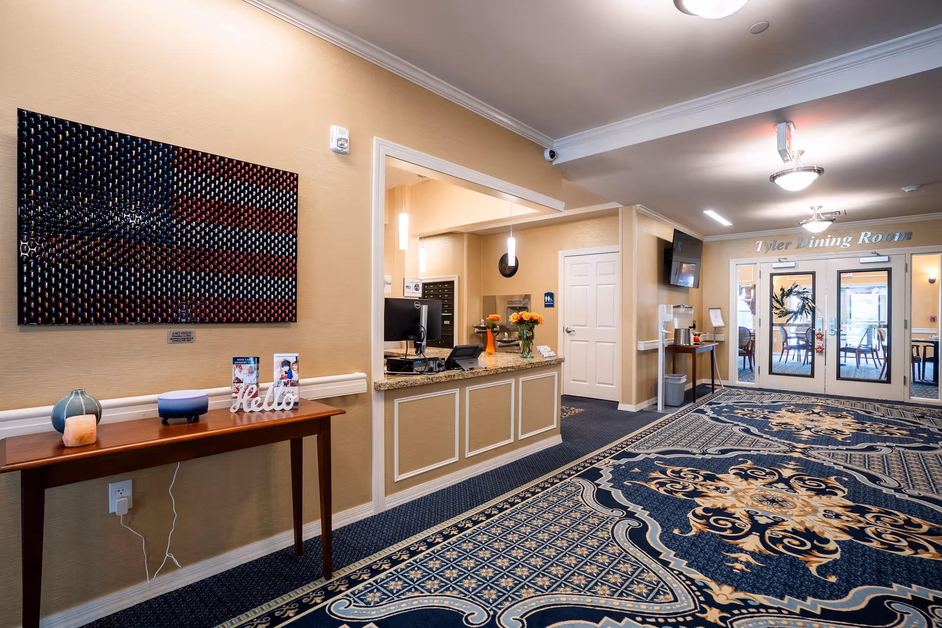 Interior view of a senior living facility reception area with a wooden table holding decorative items and brochures, a reception desk with a computer and flowers, and double glass doors leading to the Tyler Dining Room. The floor is covered with a patterned blue and beige carpet, and the walls are painted beige with crown molding.