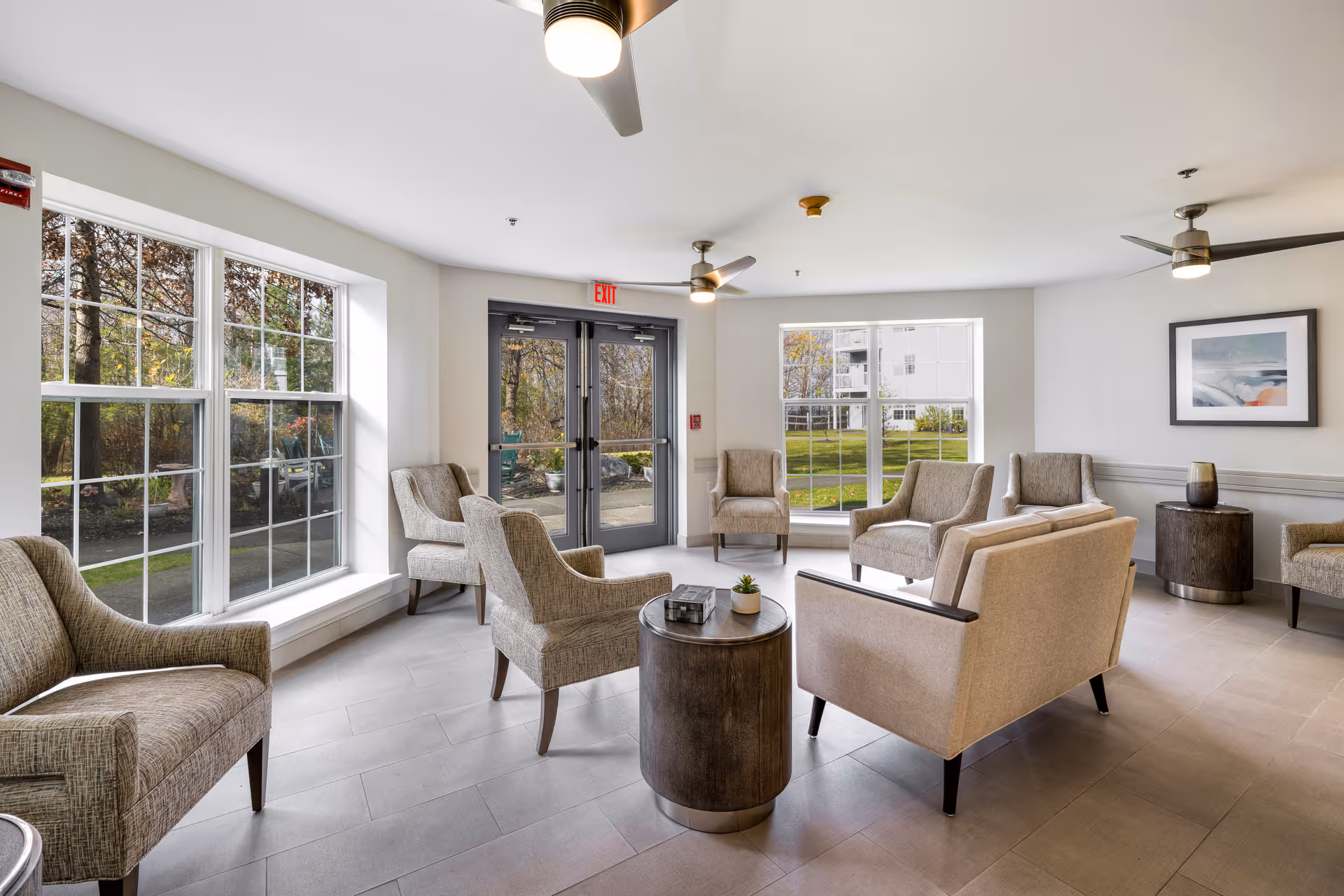A bright and spacious senior living community lounge area with multiple beige upholstered armchairs and sofas arranged around two round wooden coffee tables. Large windows and glass doors let in natural light and offer views of greenery outside. The room features ceiling fans, light gray tiled flooring, and a framed artwork on the wall.