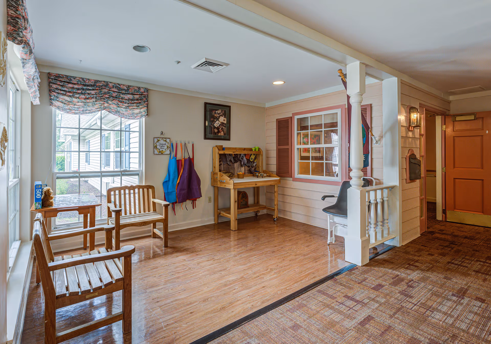 A cozy interior corner of Arden Courts - ProMedica Memory Care Community with wooden benches near large windows, colorful aprons hanging on hooks, a small wooden workbench, and a chairlift attached to a railing. The space has wood flooring and a carpeted hallway leading to a closed orange door.