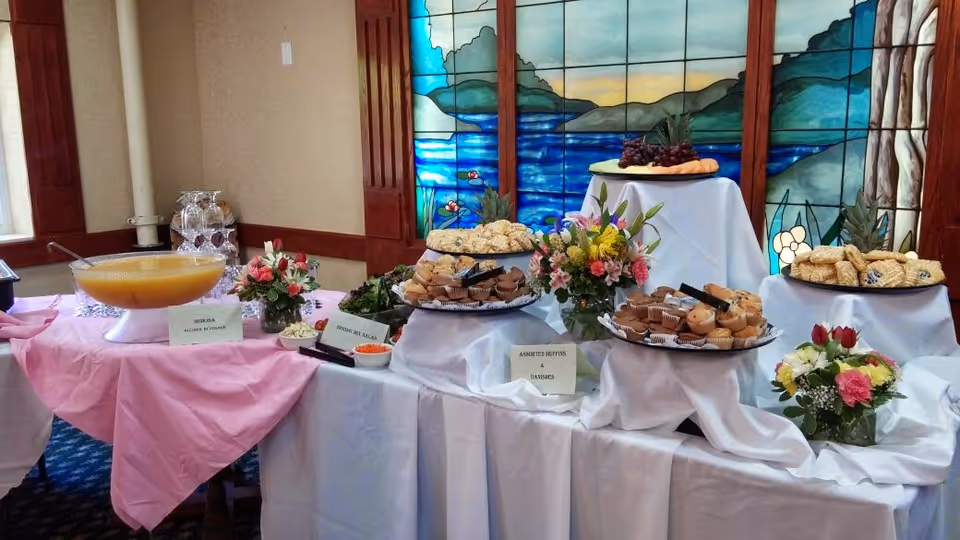 A buffet table set with various pastries, muffins, and fruit platters, decorated with floral arrangements and covered with white and pink tablecloths. The background features a large stained glass window depicting a scenic landscape with water and mountains.