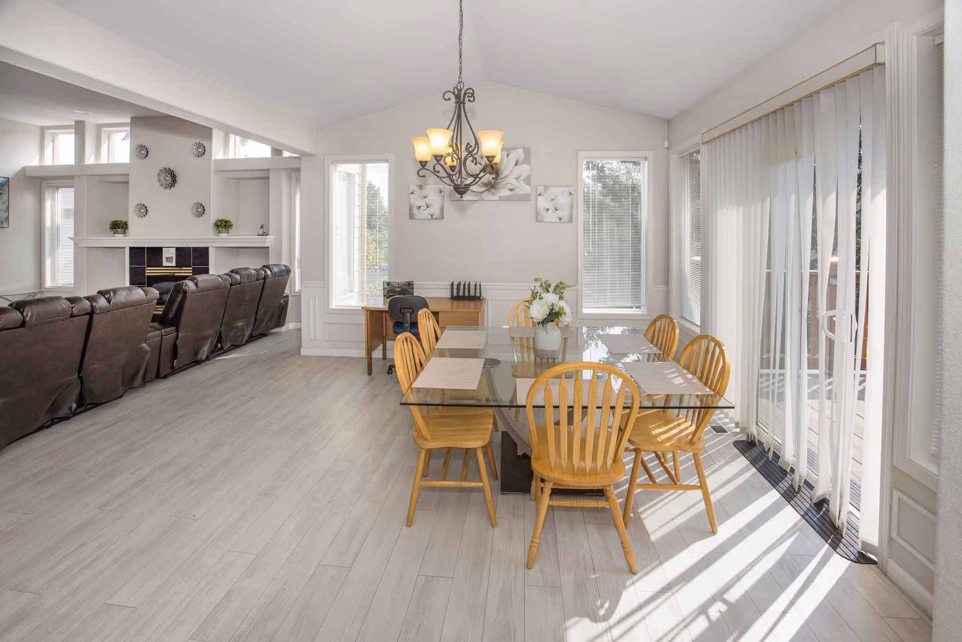 Bright dining area with a glass-top table and six wooden chairs next to sliding glass doors and an adjoining living room with leather recliners.