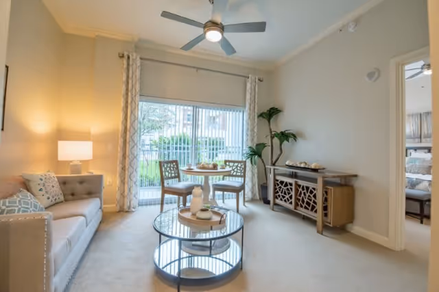 A bright living room with a beige sofa adorned with patterned cushions on the left, a round glass coffee table in the center, and a small round dining table with two chairs near a large sliding glass door with vertical blinds. A ceiling fan with a light is mounted above, and a decorative wooden sideboard with plants and ornaments is against the right wall. A doorway on the right leads to a bedroom.