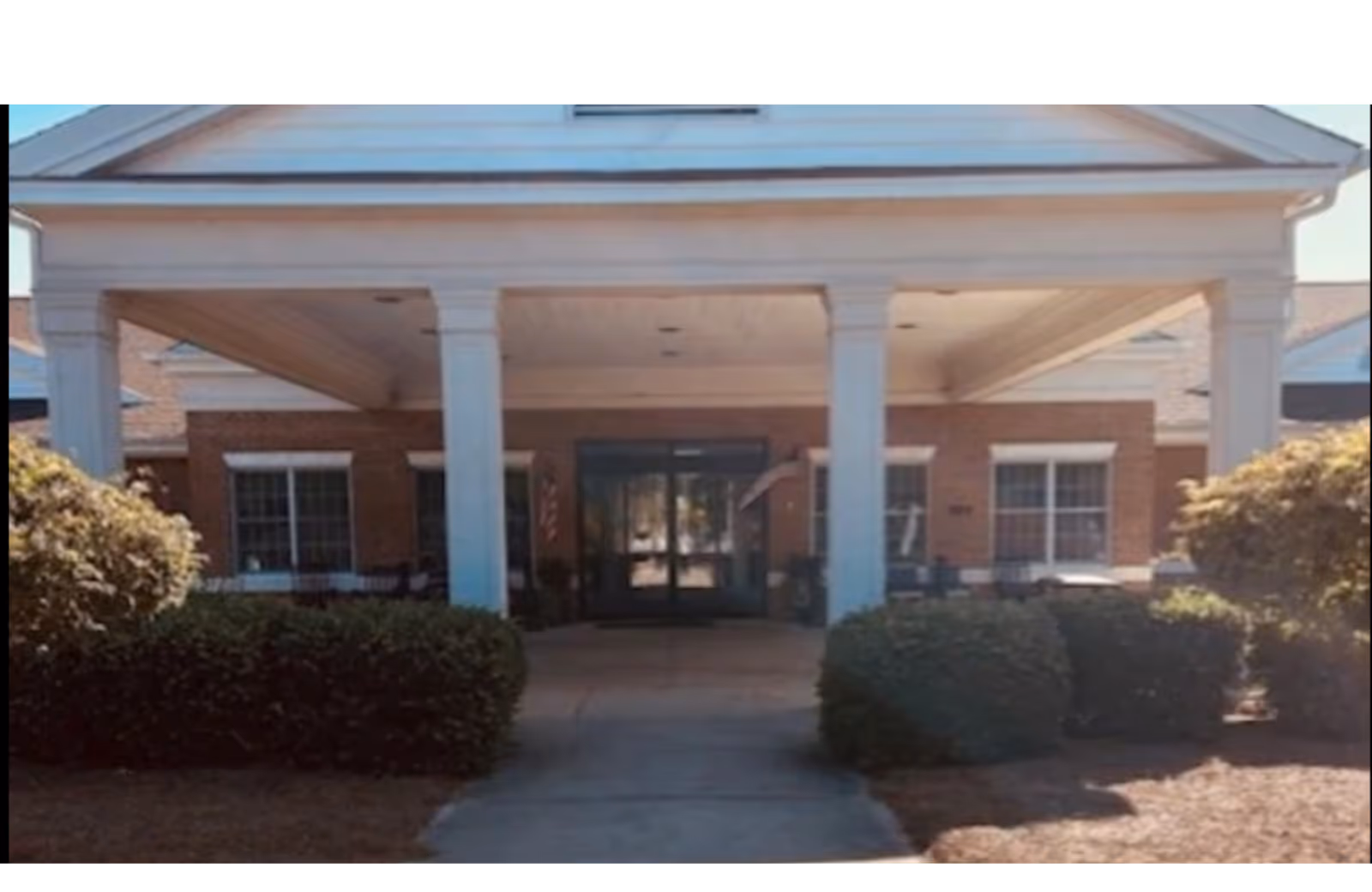 Front entrance of The Oaks Post Acute with a covered portico, white columns, double glass doors and surrounding shrubs.