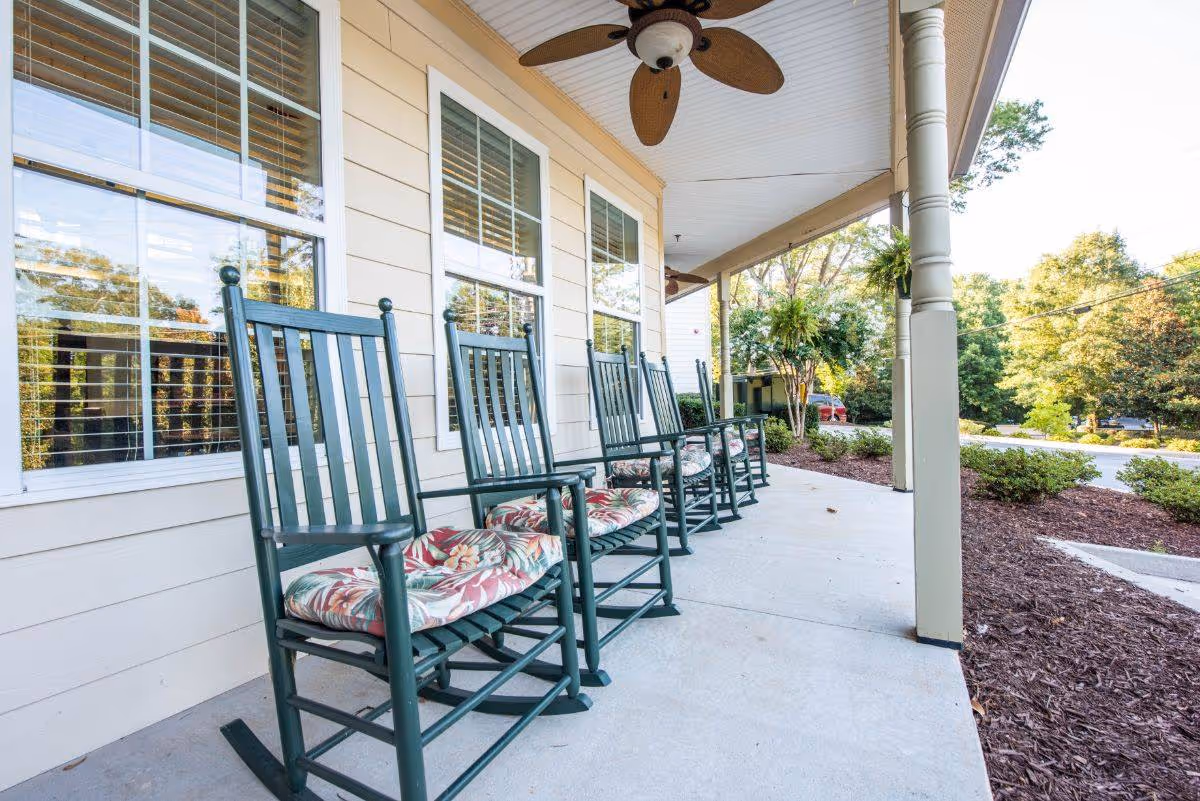 A row of green wooden rocking chairs with floral cushions lined up on a covered porch outside a building. The porch has ceiling fans and overlooks a landscaped area with bushes and trees under a clear sky.