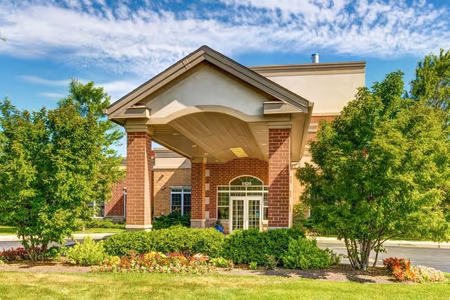 Front exterior view of Montclair Assisted Living and Memory Care building with a covered entrance, brick pillars, glass double doors, surrounded by green trees and landscaped bushes and flowers under a partly cloudy sky.