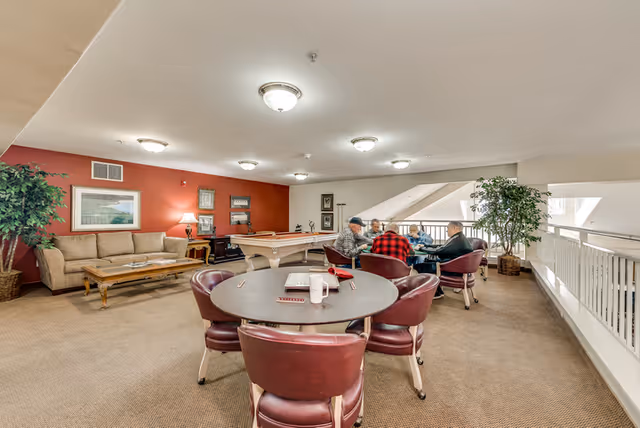 A spacious senior living community room with round card tables, a pool table, sofas, plants, and residents seated playing cards.