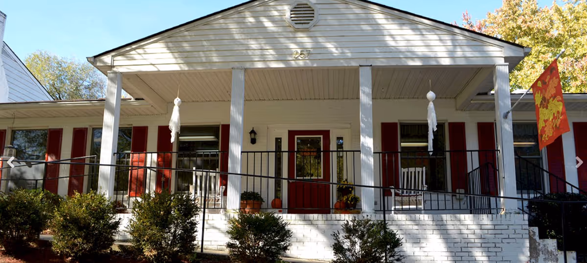 Front exterior view of a single-story building with white siding and red shutters. The entrance has a covered porch with white columns, a red door, two rocking chairs, and a black railing. There are bushes in front of the porch and a colorful flag on the right side. The building number 257 is visible above the porch.