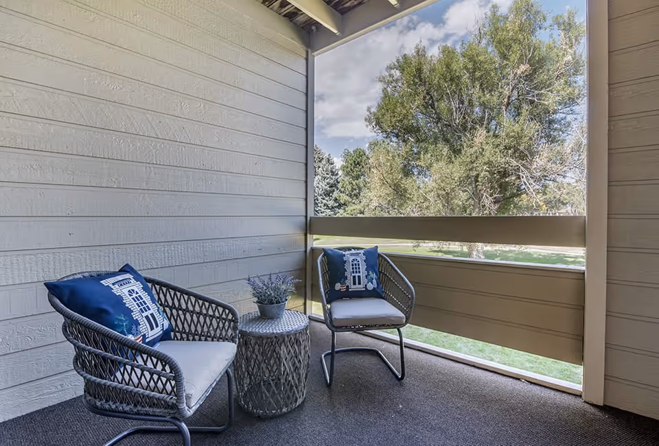 A covered outdoor balcony area with two gray woven chairs featuring blue decorative pillows and a small round table with a potted plant. The balcony overlooks a green lawn with trees under a partly cloudy sky.