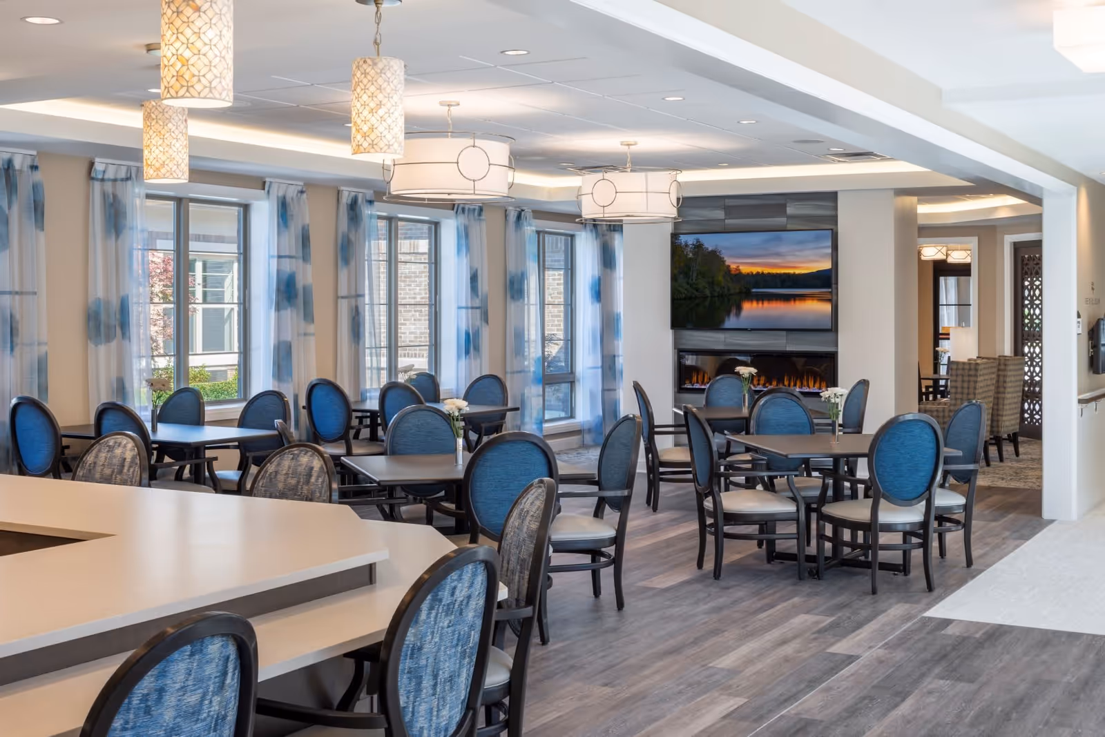 Spacious dining room with round tables, blue-upholstered chairs, pendant lights, and a wall-mounted TV above a fireplace.