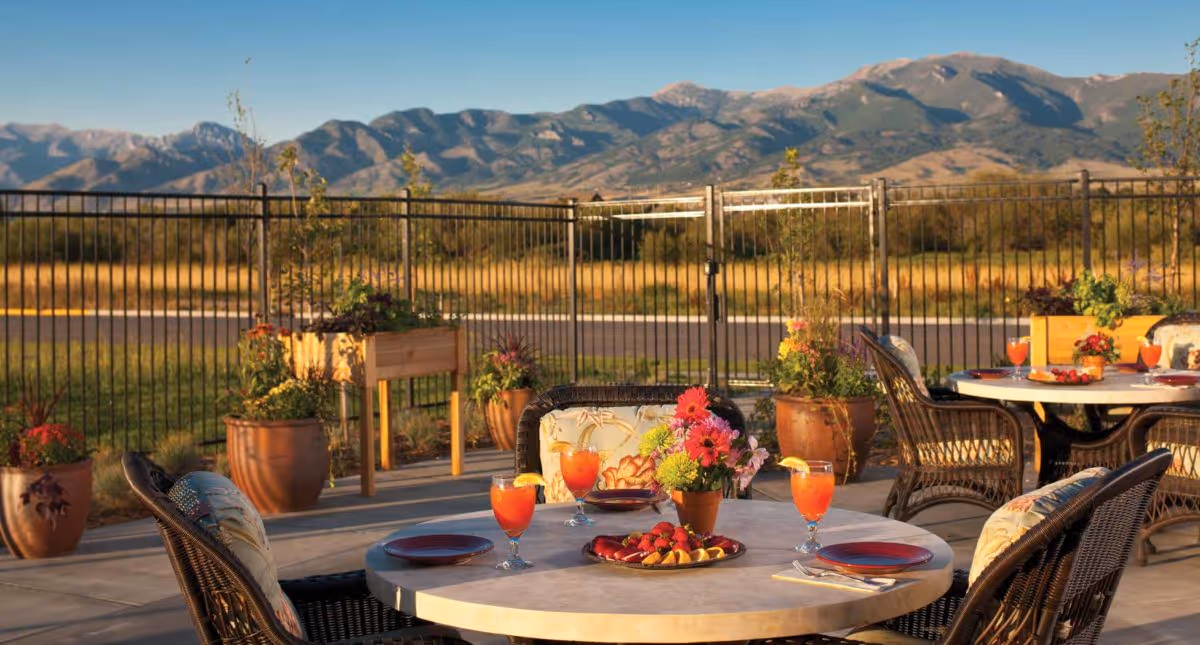 Outdoor patio area with round tables and wicker chairs, each table set with plates, glasses of orange beverage with lemon slices, and a centerpiece of colorful flowers. The patio is surrounded by potted plants and raised garden beds, with a metal fence and scenic mountain range in the background under a clear blue sky.