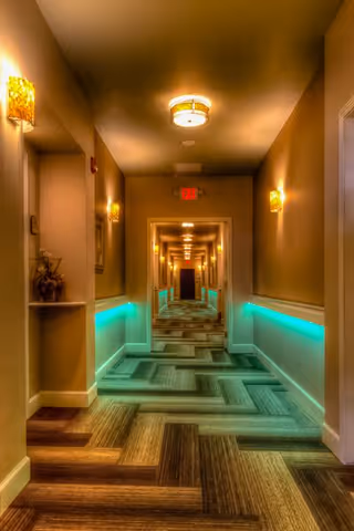 A long, well-lit hallway with patterned carpet and warm beige walls. The hallway features wall sconces and ceiling lights, with a strip of teal lighting along the lower part of the walls. There are doors along the hallway and an exit sign at the far end.