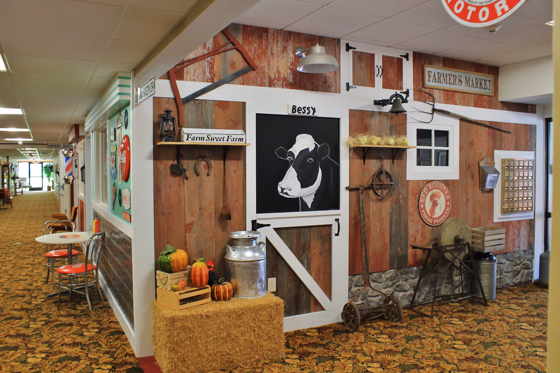 Indoor hallway corner decorated with a rustic farm theme, featuring a barn door with a painted cow labeled 'Bessy', hay bales with pumpkins, vintage farm tools, and signs such as 'Farm Sweet Farm' and 'Farmer's Market'. There are also small tables and chairs along the hallway.