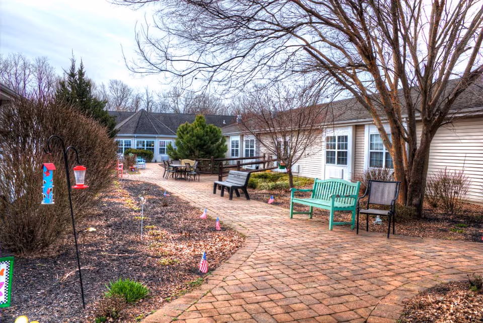Outdoor courtyard area at Merion Gardens Assisted Living featuring a brick pathway lined with benches, chairs, small American flags, bird feeders, and leafless trees and bushes. The building with multiple windows surrounds the courtyard.