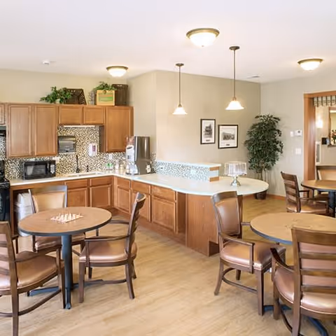Communal dining area with round tables and chairs facing a kitchenette and serving counter with pendant lights and wood cabinetry.